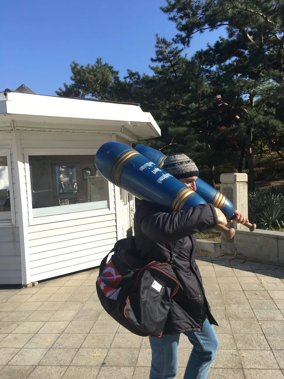 털모자를 쓴 사람이 큰 파란 페르시안밀 두 개를 양어깨에 메고 가방을 든 채 서 있다, 뒤에 소나무