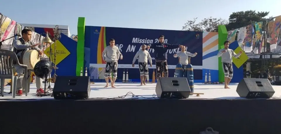 야외 무대에서 흰 상의에 수놓은 바지를 입은 여섯 명이 팔을 벌리고 움직이고, 왼쪽에서 한 사람이 북을 친다