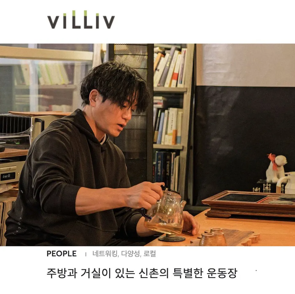 책장 앞 나무 탁자에서 검은 후드 차림의 사람이 유리 주전자로 찻잔에 차를 따른다
