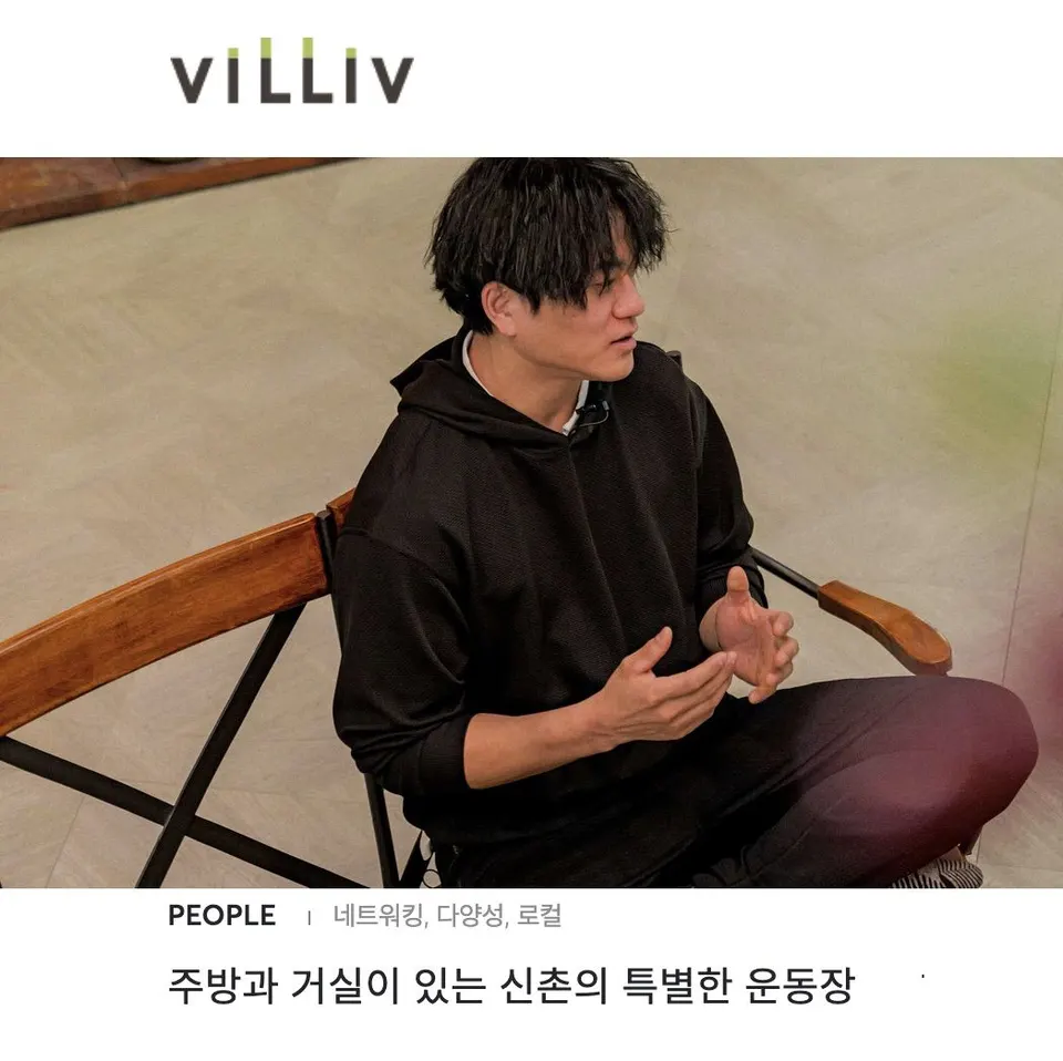 검은 후드 차림의 사람이 나무 의자 위에 책상다리로 앉아 두 손을 펴 보이며 이야기한다