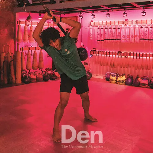 붉은 조명의 운동실에서 메이스벨을 어깨 뒤로 넘겨 휘두르는 사람, 아래에 「Den」 잡지 이름