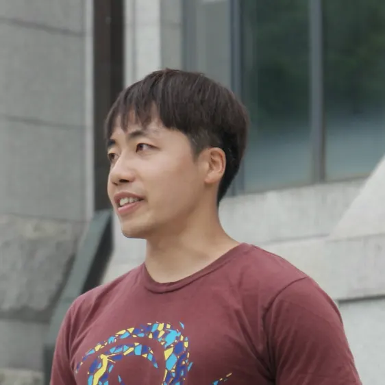돌 벽 건물 앞에서 옆을 보며 웃는 짧은 머리 사람, 자줏빛 티셔츠 차림