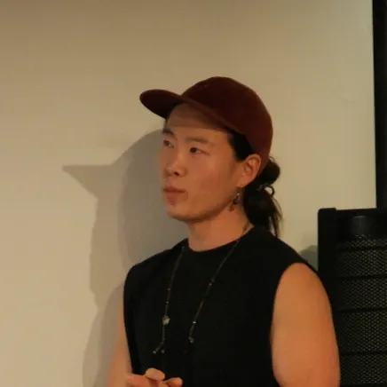 붉은 모자에 검은 민소매 차림으로 흰 벽 앞에서 옆을 바라보는 사람