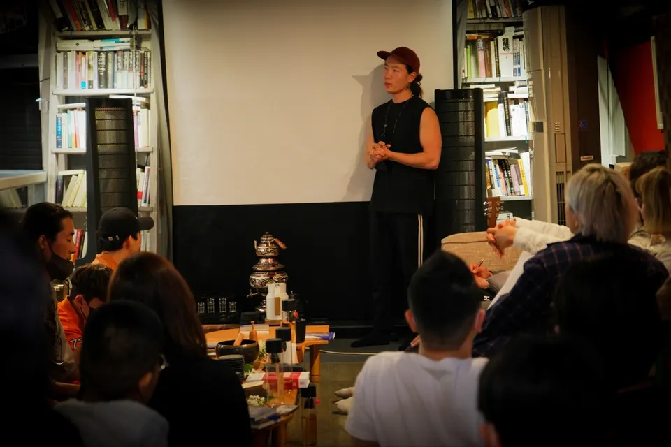 책장 사이 흰 스크린 앞에 선 사람을 바닥에 둘러앉은 참가자들이 바라본다, 앞 탁자엔 금빛 찻주전자