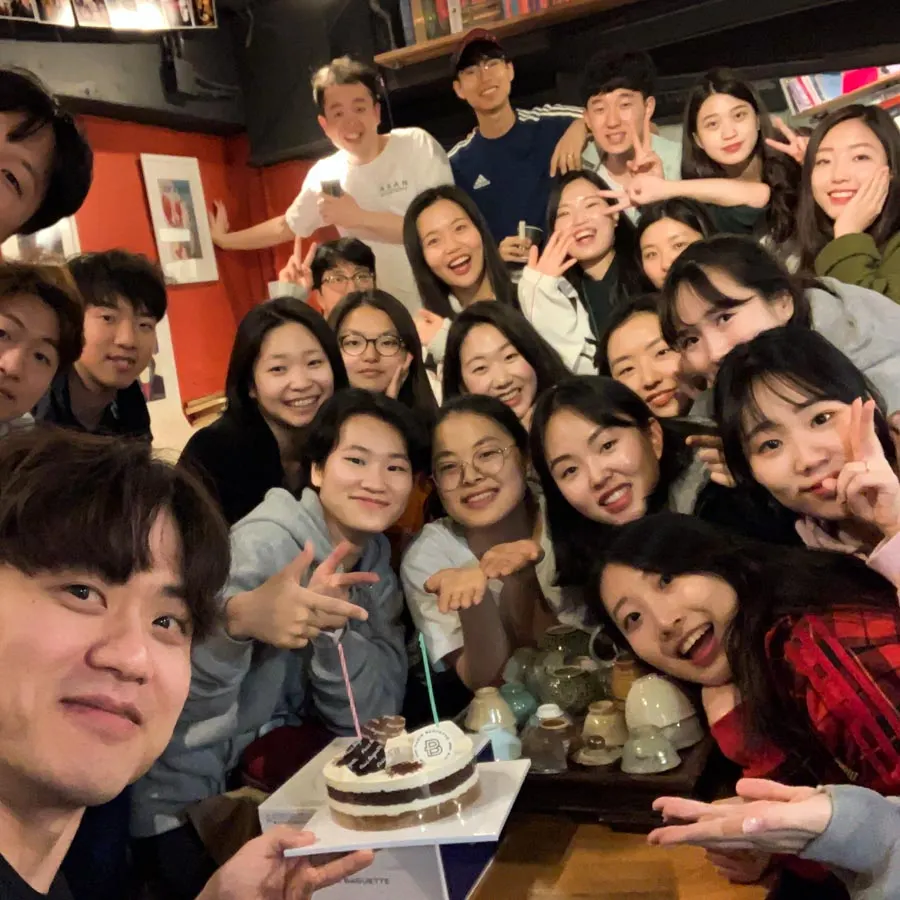 촛불을 꽂은 케이크를 앞에 두고 스무 명 남짓이 모여 브이를 하며 웃는 셀카 단체 사진