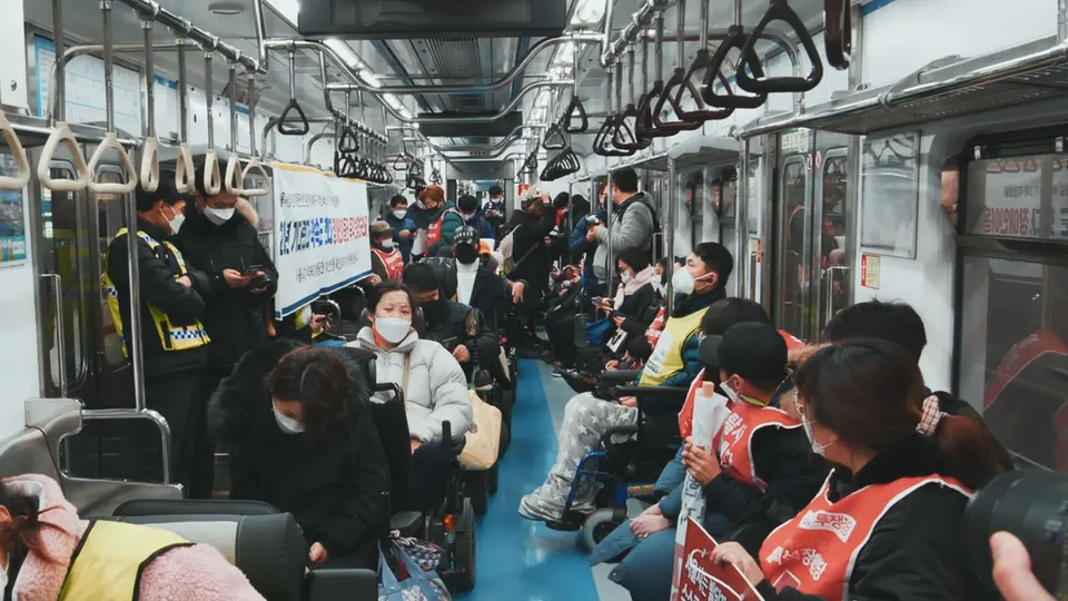 지하철 객차 안에 휠체어를 탄 사람들과 붉은 조끼를 입은 사람들이 가득하고 손팻말이 보인다