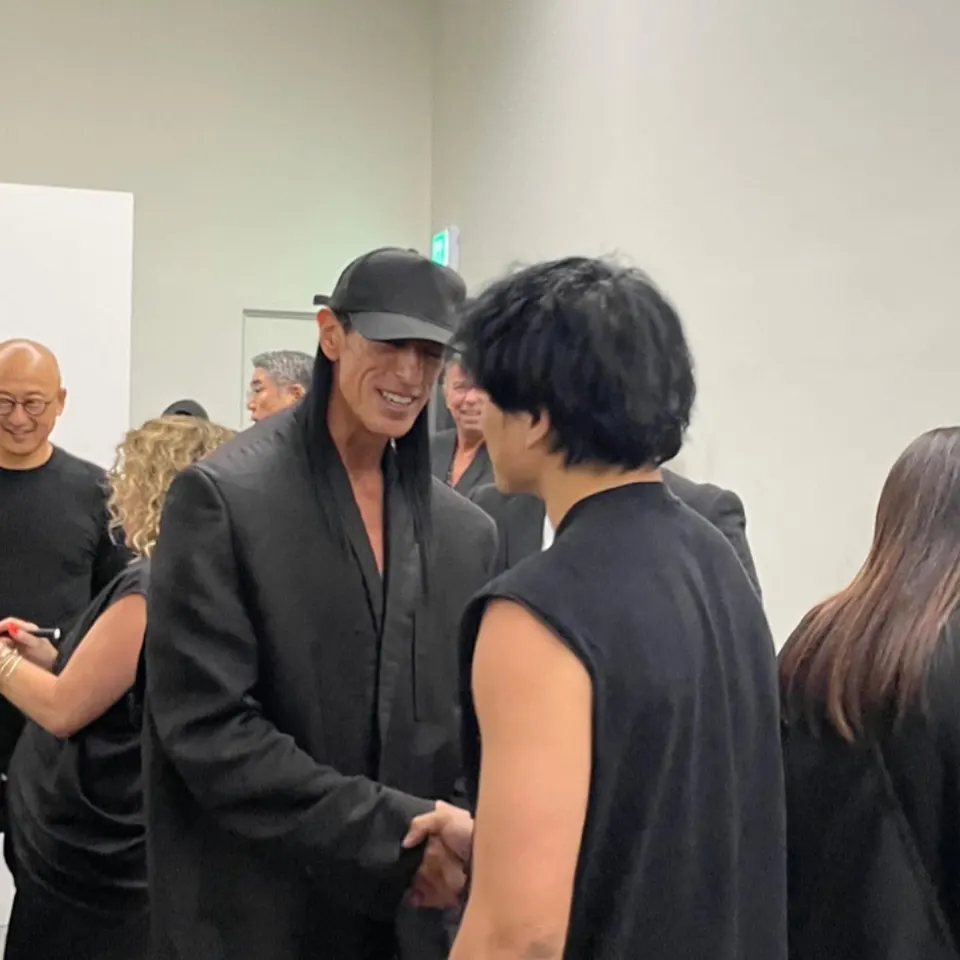 검은 모자를 쓴 사람과 민소매 차림의 사람이 웃으며 악수하고, 둘레에 사람들이 서 있다