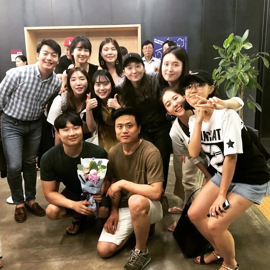꽃다발을 든 사람을 가운데 두고 열댓 명이 엄지를 들거나 브이를 하며 웃는 단체 사진