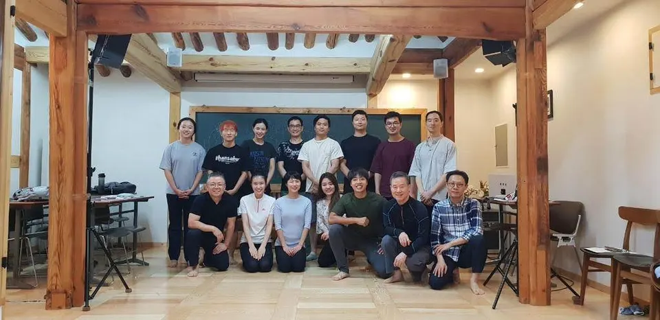 칠판과 서까래가 보이는 나무 방에서 열다섯 명이 앞줄은 앉고 뒷줄은 서서 모였다