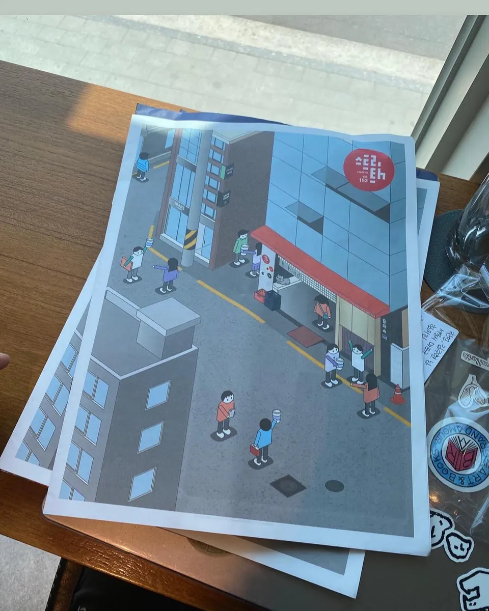 나무 탁자 위에 놓인 잡지 표지, 빨간 차양 가게가 있는 거리와 사람들을 그린 그림