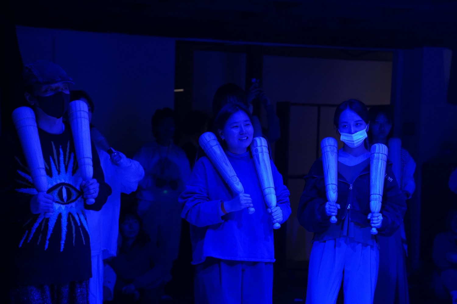 세 사람이 밝은 나무 방망이 한 쌍씩을 가슴 앞에 세워 들고 서 있다, 둘은 마스크를 썼다