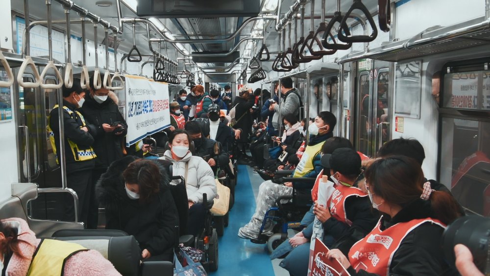 지하철 객차 안에 휠체어를 탄 사람들과 붉은 조끼를 입은 사람들이 가득하고 손팻말이 보인다